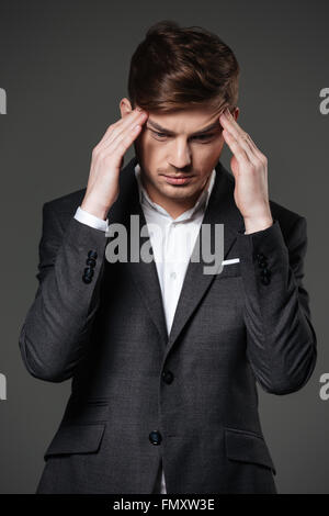 Upset unhappy young businessman in black suit touching his temples and having a headache over grey background Stock Photo