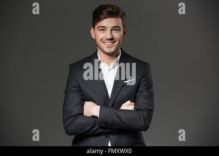 Smiling attractive young businessman in black suit standing with hands folded  over grey background Stock Photo