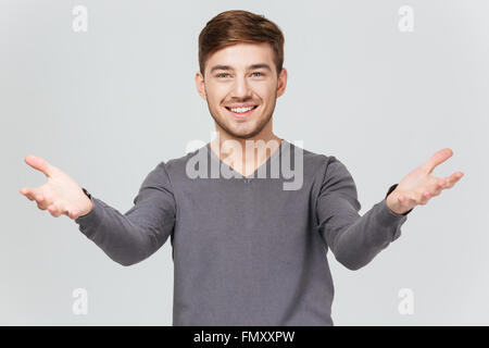 Cheerful handsome young man in grey pullover smiling and showing welcoming gesture over white background Stock Photo