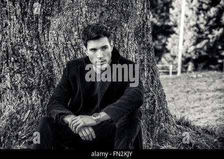 Handsome young man leaning against tree, looking to a side, in a sunny day wearing a black coat Stock Photo