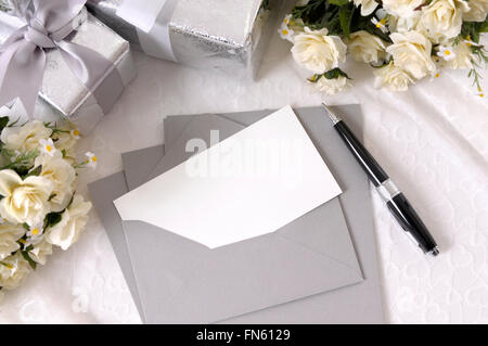 Writing paper or wedding invitation with envelope laid on bridal lace with several wedding gifts and white rose bouquets.  Space Stock Photo