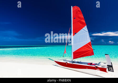 Sailing boat with red sail on a beach of deserted tropical island with shallow blue water Stock Photo