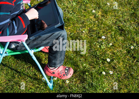 A young child using an ipad while sat outside on a  small deck chair.