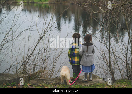 Boy, girl and golden retriever puppy dog standing at water's edge Stock Photo