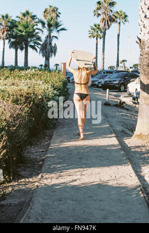 A woman in a black bikini walking down a path carrying a surfboard on her head. Stock Photo