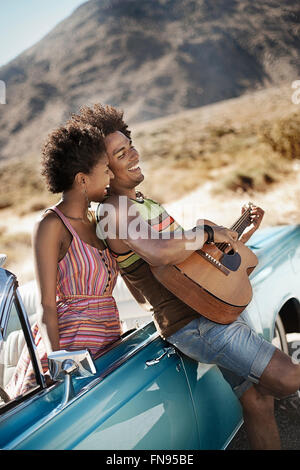 A young couple standing by a pale blue convertible on the open road, the man playing a guitar. Stock Photo