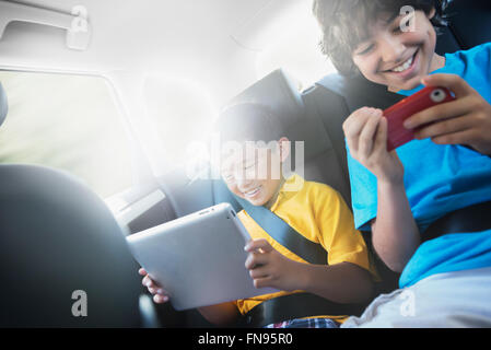 Two children travelling in the back seat of a car, one using a digital tablet and one a handheld game. Stock Photo