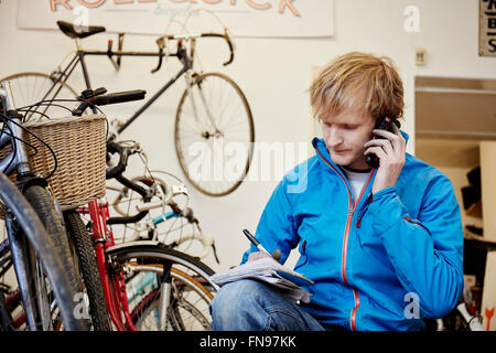 A young man using his smart phone in a cycle shop, making a call. Stock Photo