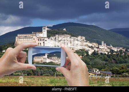 In the left bottom of the photo are hands holding smart phone and taking picture of Assisi town in Umbria (Italy) Stock Photo