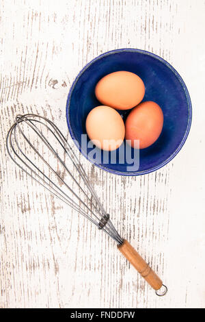 Three eggs in a ceramic bowl with an old wisk on a rustic painted wood table top shot from above. Stock Photo