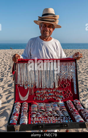 A Mexican man wears many hats for sale and sells silver jewelry souvenirs on the beach at San Jose del Cabo, Los Cabos, Mexico. Stock Photo