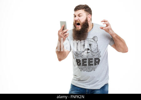 Irritated angry man with beard holding smartphone and shouting over white background Stock Photo