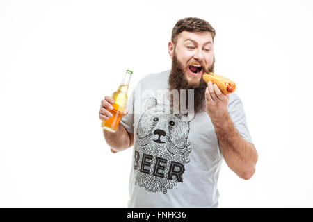 Handsome bearded man holding bottle of beer and eating hot dog over white background Stock Photo