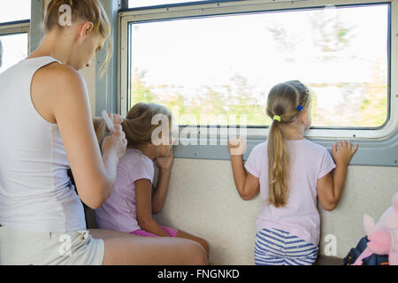 two girls looking out of the window while sitting in an electric train, a young mother combing long hair of one of them Stock Photo