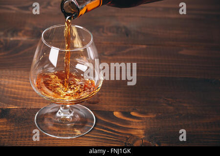 Pouring brandy or cognac from the bottle into the glass against wooden background Stock Photo