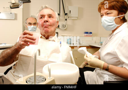 An elderly man rinses with anti-microbial mouth wash after dental treatment, watched by the dentist's assistant Stock Photo