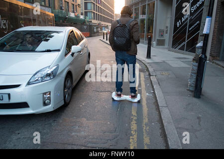 Man commuting on his hoverboard passes other traffic going in the opposite direction. London, England, UK. A self-balancing two-wheeled board, or self-balancing electric scooter, also commonly referred to as a hoverboard, is a type of portable, rechargeable battery-powered scooter. They typically consist of two wheels arranged side-by-side, with two small platforms between the wheels, on which the rider stands. The device is controlled by the rider's feet, standing on the built-in gyroscopic, sensored pads. Stock Photo