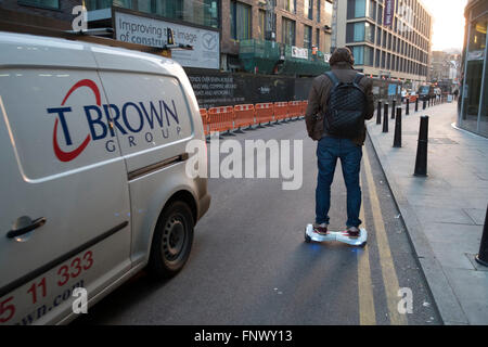 Man commuting on his hoverboard passes other traffic going in the opposite direction. London, England, UK. A self-balancing two-wheeled board, or self-balancing electric scooter, also commonly referred to as a hoverboard, is a type of portable, rechargeable battery-powered scooter. They typically consist of two wheels arranged side-by-side, with two small platforms between the wheels, on which the rider stands. The device is controlled by the rider's feet, standing on the built-in gyroscopic, sensored pads. Stock Photo