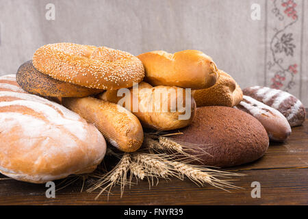 Pile of different types of baked bread loaves, baguettes, bagels and rolls on top of whole wheat stalks over wood table Stock Photo