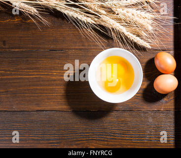 Fresh eggs broken into a bowl with ears of wheat and two extra eggs on a wooden table with copy space viewed from above conceptual of baking. Stock Photo