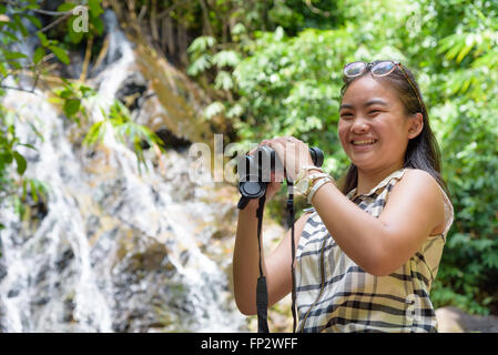Beautiful young girl hiking is using binoculars look for birds smiling to the camera in tropical forest near the waterfalls Stock Photo