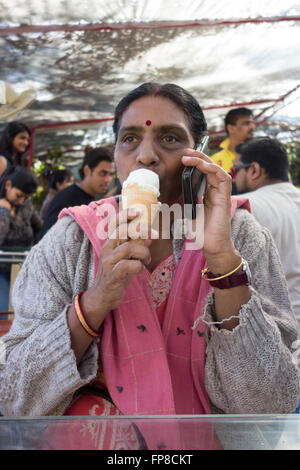 Busy Indian woman eating vanilla ice cream cone, holding mobile phone. Stock Photo