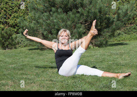 Front view of flexible blonde senior woman is stretching exercising outdoors. The smiling woman is looking at the camera. Stock Photo