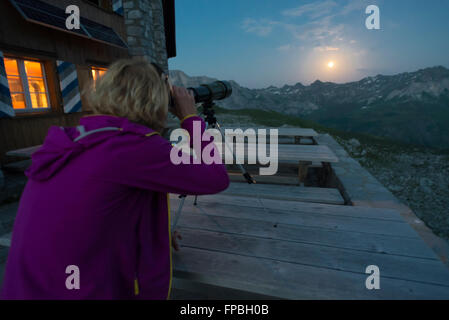 A woman looks at the full moon over Prättigau mountains with a telescope on the Carschina mountain hut in the Swiss Alps,Grisons Stock Photo