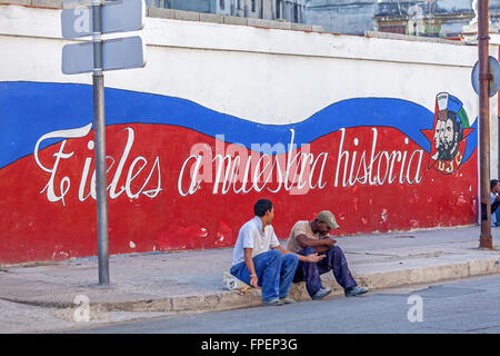 HAVANA, CUBA - APRIL 2, 2012: Two cuban teenagers sitting near propaganda graffiti Stock Photo