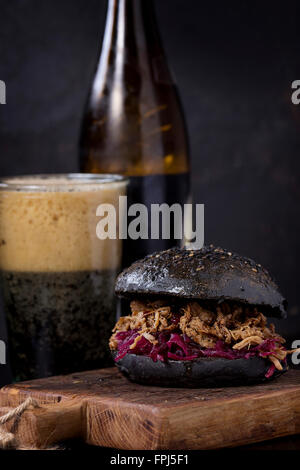 Black homemade burger with beef stews and red cabbage served on small wooden chopping board with glass and bottle of dark beer o Stock Photo