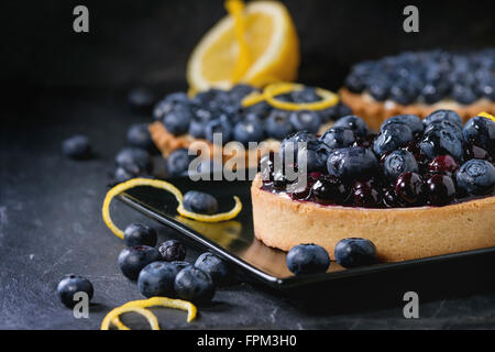 Lemon Tart and tartlets with fresh and cooked blueberries, served on black square plate with lemon and lemon zest over black bac Stock Photo