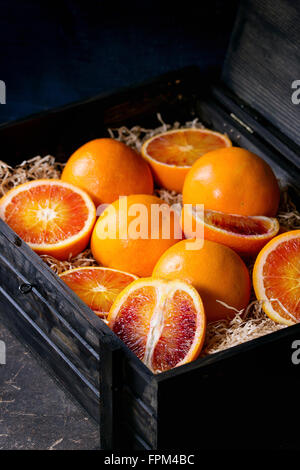 Sliced and whole Sicilian Blood oranges fruits in black wooden box with sawdust over dark background. Stock Photo