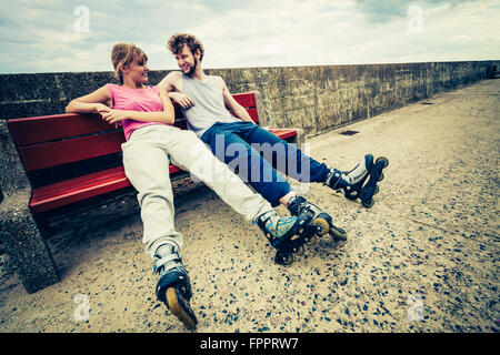 Young people friends in training suit with roller skates. Woman and man relaxing on bench outdoor. Stock Photo