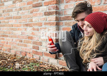 Young couple listening to music on mobile phone against brick wall, Munich, Bavaria, Germany Stock Photo