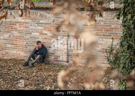 Young man talking on mobile phone and sitting on skateboard leaning against brick wall, Munich, Bavaria, Germany Stock Photo