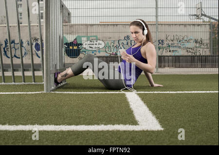 Young woman taking rest after exercising and listening to music on football ground, Bavaria, Germany Stock Photo