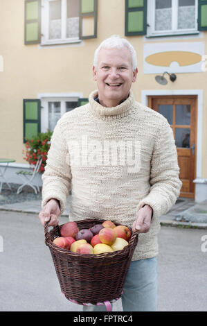 Portrait of a mature man holding basket full of apples in his hands in front of wholefood shop and smiling, Bavaria, Germany Stock Photo