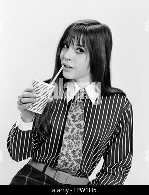 1960s YOUNG WOMAN MOD FASHION DRINKING FROM PAPER CUP STRAW STRIPED SHIRT PRINT NECKTIE LOOKING AT CAMERA Stock Photo