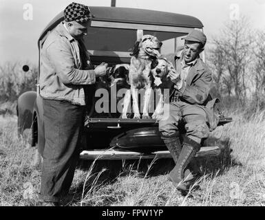 1930s TWO MEN IN HUNTING CLOTHES SITTING STANDING WITH ENGLISH SETTER DOGS ON BACK OF WOODIE STATION WAGON Stock Photo