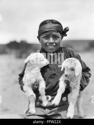 1930s SMILING NATIVE AMERICAN NAVAJO BOY LOOKING AT CAMERA HOLDING TWO BABY LAMBS NEW MEXICO USA Stock Photo