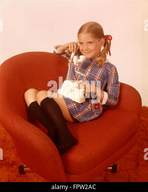 1970s SMILING GIRL KNEE SOCKS PLAID DRESS SITTING RED CHAIR TALKING PHONE TELEPHONE Stock Photo