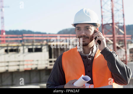 Young male engineer holding blueprint and talking on a mobile phone at construction site, , Baden-Württemberg, Germany Stock Photo