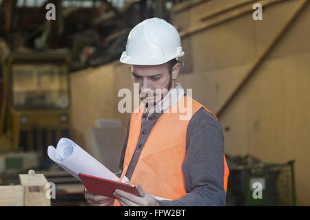 Young male engineer holding blueprint and using digital tablet at construction site, , Baden-Württemberg, Germany Stock Photo