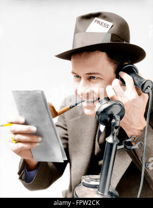 1930s EXCITED MAN NEWSPAPER REPORTER SMOKING PIPE PRESS PASS IN HAT TALKING ON UPRIGHT PHONE REPORTING STORY FROM NOTE PAD Stock Photo
