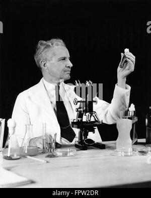 1940s SENIOR MAN SCIENTIST CHEMIST HOLDING UP TEXT TUBE WORKING IN LAB MICROSCOPE VIALS BEAKERS RESEARCH Stock Photo