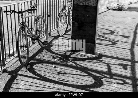 Two road city bikes locked to a metal fence. next to a road. Big shadows projected on the pavement.Black and white. Stock Photo