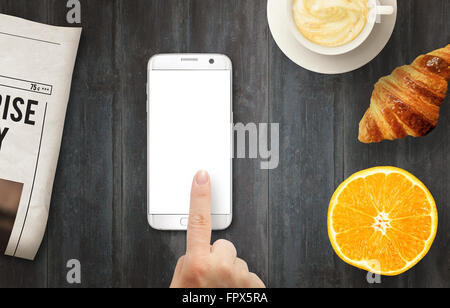 Smart phone with isolated display for mockup. Hand touching display. Newspapers, coffee, croissant and orange on table. Top view Stock Photo