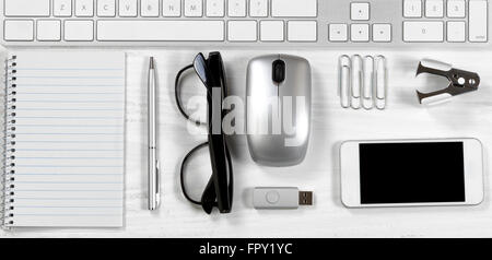 Overhead view of a white desktop with basic office stationery objects lined up under keyboard. Stock Photo