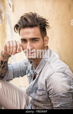 Portrait of handsome man with tattoo on arm sitting on cement stairs while leaning on wall, looking at camera Stock Photo