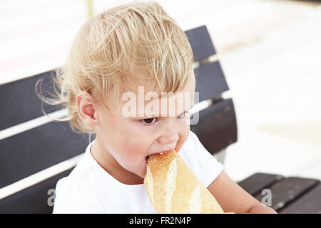 Outdoor close up portrait of cute Caucasian blond baby girl eating big French baguette Stock Photo